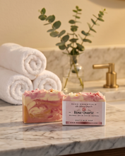 Two decorative handmade soap bars with pink and cream swirls sit on a marble countertop beside rolled white towels and a glass vase with green stems. The front soap bar has a label reading 'Redo Essentials Rose Quartz' and 'Coated Milk Soap'.