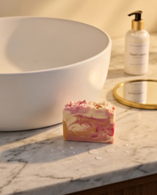 A rectangular handmade soap bar with pink and cream marbled swirls sits on a marble bathroom counter next to a white ceramic sink, with a hand lotion bottle and gold mirror in the background.