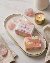 A styled product photo of a handmade soap bar and its packaging on a white oval tray with gold trim, surrounded by rose quartz crystals, a lit candle, and neutral textiles on a marble surface.