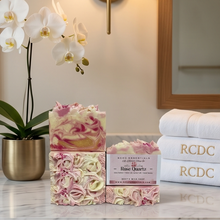Two decorative handmade soap bars labeled 'Rose Quartz' with pink and cream swirls and layered rose petals, displayed on a marble countertop in a bathroom setting with a potted orchid, wall sconce, and folded white towels.