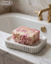 A rectangular bar of soap with pink and cream swirls sits on a white marble soap dish on a bathroom sink. The soap has coarse salt crystals on top, with a blurred gold faucet and tiled wall in the background.