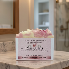 A boxed bar of goat's milk soap labeled 'Rose Quartz' by RCDC Essentials sits on a marble bathroom counter. The soap top is swirled pink and white, and the background shows a softly blurred bathroom interior with wood trim and a mirror.
