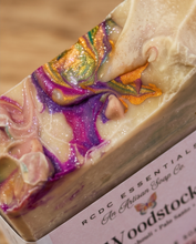 A close-up of a rectangular bar of handmade soap with a marbled swirl pattern in cream, purple, pink, green, and orange, with glitter. The soap bar is shown with its paper label from 'RCDC Essentials' partially visible.