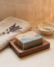 A rectangular bar of marbled soap with pastel swirls sits on a dark wooden soap dish on a beige countertop. A folded beige cloth with dried lavender sprigs rests beside it, and a small speckled bowl is blurred in the background against warm beige tiles.