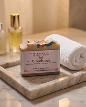A bar of RCDC Essentials Woodstock goat's milk soap sits on a marble tray next to a rolled white towel and two perfume bottles in a warm bathroom setting.