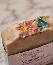 A close-up of a handmade milk soap bar in a paper sleeve, featuring a beige base with colorful marbled swirls of gold, pink, purple, and teal. The label reads 'RCDC ESSENTIALS As Artisan Soap Co.