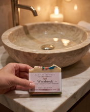 A hand holds a bar of goat's milk soap labeled 'Woodstock' in front of a warm, softly lit bathroom scene with a stone vessel sink and a candle.