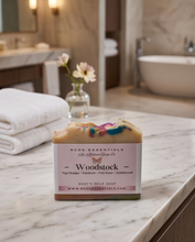 A bar of handmade soap labeled 'Woodstock' by 'MCDC Essentials' sits on a white marble bathroom counter, with folded white towels and a small vase of flowers blurred in the background.