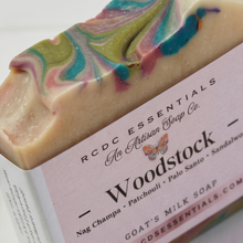 A close-up of a handmade milk soap bar with a pink label. The soap has a creamy beige base with colorful swirls of green, blue, and pink.