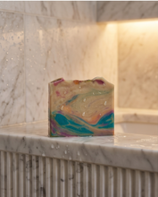 A handmade soap bar with a marbled pattern of teal, orange, purple, and cream sits on a wet white marble countertop in a bathroom setting.