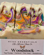 A close-up of a handcrafted soap bar with three vertical cuts, showing a marbled swirl pattern of cream, purple, orange, green, and pink with glitter. White dripping wax sits on top, and a paper label at the bottom reads 'RCDC ESSENTIALS An Artisan Soap Co.