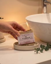 A hand holds a bar of RCDC Essentials Woodstock handmade soap over a small dish in a warm, modern bathroom setting with a white vessel sink and eucalyptus leaves.