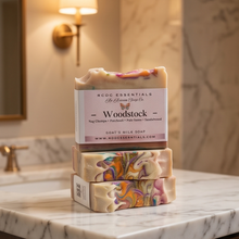 Three stacked bars of NCCO Essentials Woodstock Goat's Milk Soap on a white marble bathroom counter, with a warm wall sconce and marble wall in the background.