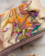 A close-up of a rectangular bar of soap with a marbled swirl pattern in cream, orange, purple, green, blue, and pink, resting on a light marble surface.