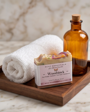 A bar of handmade soap labeled 'Woodstock' by RCOC Essentials sits on a wooden tray next to a rolled white towel and an amber glass bottle, set against a marble background.