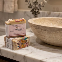 Three stacked bars of handmade milk soap with colorful swirls and a pink label reading 'Woodstock' and 'MCC Essentials by Natural Soap Co,' displayed on a white marble countertop beside a large stone bowl in a bathroom setting.