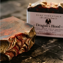 A close-up of a decorative bar of soap with a textured, leaf-like pattern in metallic gold and deep red, shown next to its white paper packaging box labeled 'Dragon's Blood' by MCGG Essentials.
