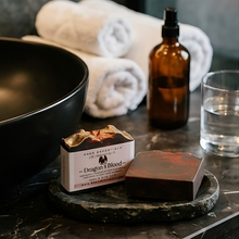 A bar of soap labeled 'Dragon's Blood' by Mark DeGrootse & Co. sits on a dark stone tray on a marble countertop, next to a glass of water, an amber spray bottle, and rolled white towels in the background.