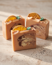 Three decorative bars of soap with orange slice garnishes and small green leaf accents, arranged on a light wood surface with a few scattered black seeds.