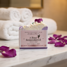A rectangular bar of soap labeled 'A Rose Remembered' by RCOC Essentials, styled on a white marble surface with white rolled towels and purple rose petals in the background.