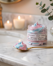 A jar of whipped soap scrub labeled 'Mermaid Dreams' sits on a white marble surface with a gold spoon holding a swirl of pink and blue scrub in front. The background is softly blurred with lit candles and greenery.
