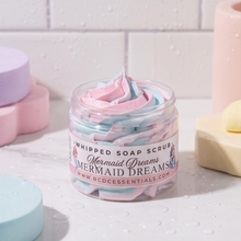 A clear jar of whipped soap scrub labeled 'Mermaid Dreams' by RCO Cessentials, filled with pink and blue whipped lather, set on a wet white surface with pastel bath items around it.
