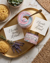 A styled flat-lay of a spa gift set on a gold oval tray, featuring a jar of pink and blue whipped soap, a jar of 'Mermaid Dreams' whipped body scrub, two white cards reading 'Elevate Your Ritual', a gold spoon, a yellow sponge, and lavender sprigs on a marble surface.