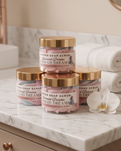 Four jars of Peony Essentials whipped soap scrub in the 'Mermaid Dreams' scent, stacked on a white marble countertop with a white orchid flower and folded white towels in the background.
