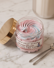 A jar of whipped soap scrub labeled 'Mermaid Dreams' sits on a marble surface with its gold lid beside it. The scrub is swirled pink and light blue, and two metal spoons are placed nearby.