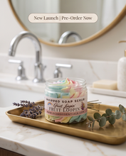 A jar of GCO Essentials Fruit Loopin Whipped Soap Scrub sits on a gold tray on a marble countertop in a bathroom setting with a sink and mirror in the background. A banner at the top reads 'New Launch | Pre-Order Now'.