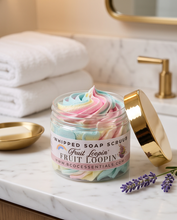 A jar of whipped soap scrub labeled Fruit Loopin' by BCC Essentials, filled with pastel pink, blue, and yellow whipped texture, set on a marble bathroom counter with a gold lid leaning beside it.