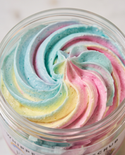 A close-up of a whipped cosmetic scrub in a clear jar, swirled with pastel blue, pink, yellow, and lavender tones and small visible specks, with partial text reading 'WHIPPLED SCRUB' on the jar.