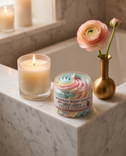 A jar of whipped soap scrub labeled 'Fruit Loops' sits on a marble bathroom counter beside a lit candle and a gold vase holding a pink flower, with a bathtub in the background.