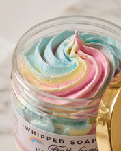 A close-up of a whipped soap in a clear glass jar with a white label and a gold lid resting beside it. The soap has a pastel swirl of blue, pink, yellow, and cream, with the text 'Whipped Soap Fruit Loop' partially visible on the label.
