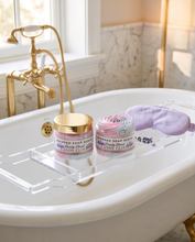 Two jars of whipped soap scents labeled 'Candy Clog' and 'Candy Clog Spice' with lavender sprigs, displayed on a clear acrylic bath tray resting on a white clawfoot bathtub. A lavender sleep mask is also present, with a gold faucet and marble wall in the background.