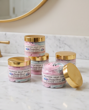 Five jars of Whipped Soap Scrub in the Candy Cloud scent, arranged on a white marble countertop with a gold-framed mirror in the background. Two jars are open, showing the whipped pink and blue scrub inside, while three are closed with shiny gold lids.