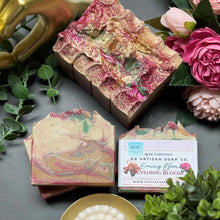 Artisan soap bars with a visible brand label, surrounded by flowers and greenery on a gray surface.